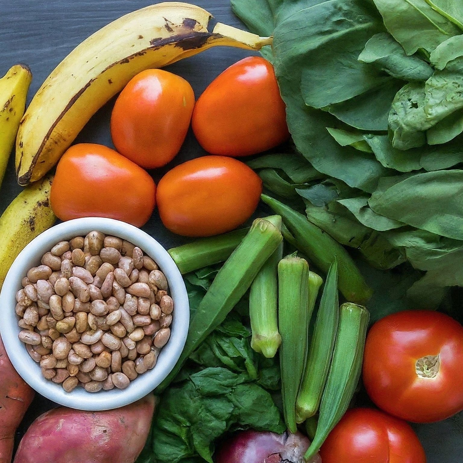 A close-up shot of fresh ingredients for Nigerian cuisine including yams, sweet potatoes, plantains, leafy greens, tomatoes, bell peppers, okra, and a bowl of beans.